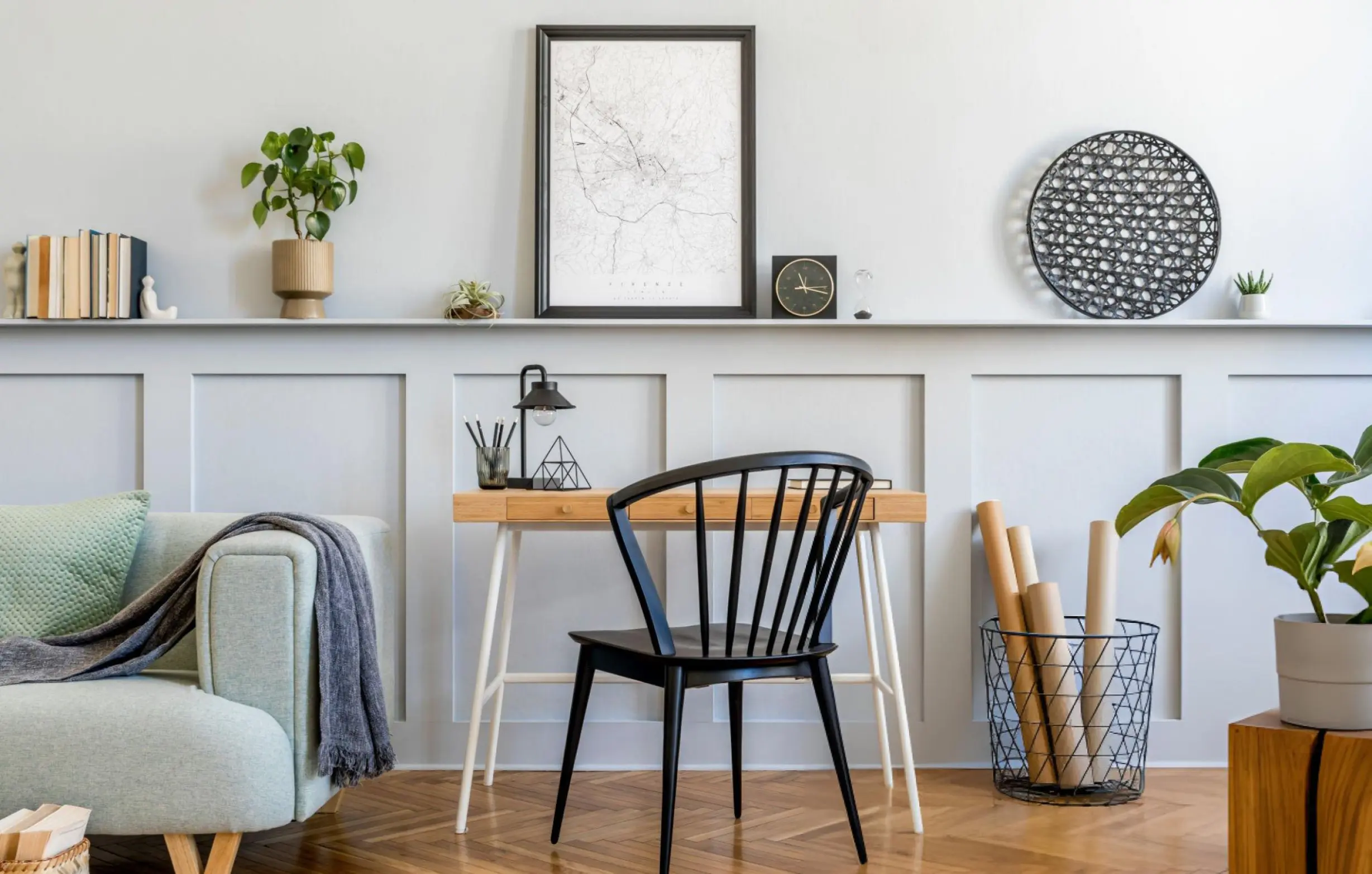 A styled interior scene with a light gray sofa, a black spindle-back chair, a wooden desk, framed wall art, plants, and home decor against a pale gray wall.